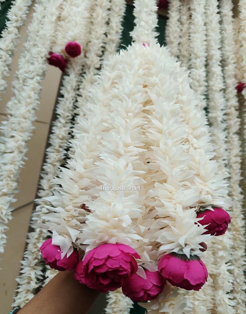 Op de afbeelding: Een close-up van een witte bloemenslinger met roze accenten. De slinger is gemaakt van gelaagde, getextureerde witte bloemblaadjes en heeft clusters van levendige roze bloemen. De slinger hangt verticaal.