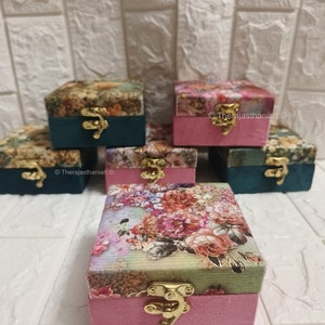 Puede incluir: Cuatro pequeñas cajas rosas con tapas de estampado floral y cierres dorados. Las cajas están dispuestas en forma de pirámide sobre una superficie de azulejos blancos.
