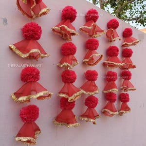 May include: Red decorative hanging ornaments with a pom-pom top and a flared skirt trimmed with gold. The ornaments are arranged in rows and columns on a light pink wall. The text "THERAJASTHANIART" is visible on the wall.