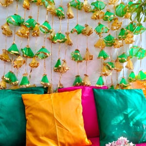 May include: A pink sofa with three cushions, one yellow, one pink and one green. The cushions are decorated with gold trim. The background is a white wall with a garland of green and gold fabric flowers hanging from the top.