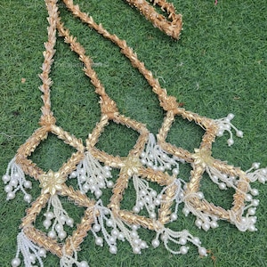 May include: Gold-colored decorative garland with a floral design and white pearl-like beads. The garland is made of woven material and is arranged on a green surface. The text "the rajasthan art" is visible.