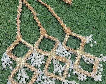 May include: Gold-coloured decorative garland with a floral design and white pearl-like beads. The garland is made of woven material and is arranged on a green surface. The text "the rajasthan art" is visible.