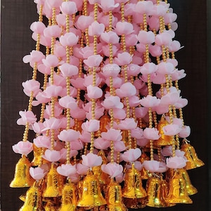 May include: A pink flower garland with gold bells hanging from it. The garland is made of fabric flowers and gold beads. The bells are ornate and have a gold finish.