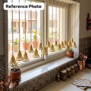 May include: A window treatment featuring multiple gold-toned metal chains hanging vertically, each ending in a small, intricate, bell-shaped lattice weight. The chains are suspended in front of a window with potted plants visible on the sill.