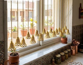 May include: A window treatment featuring multiple gold-toned metal chains hanging vertically, each ending in a small, intricate, bell-shaped lattice weight. The chains are suspended in front of a window with potted plants visible on the sill.