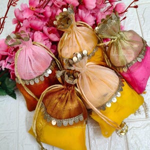 Könnte beinhalten: Eine Sammlung farbenfroher Beutel mit Kordelzug in Rosa, Orange, Gelb und Gold. Jeder Beutel ist mit metallischen Verzierungen und einem Perlenquasten versehen. Die Beutel sind mit rosa Blüten im Hintergrund angeordnet.