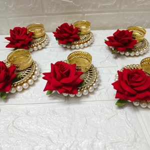 May include: Six gold candle holders with red rose embellishments and pearl accents. The candle holders are arranged in a circle on a white surface.