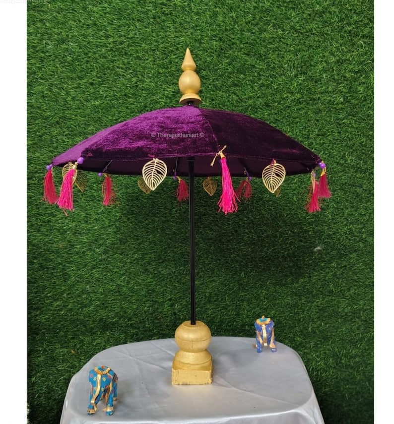 K&ouml;nnte beinhalten: Ein violetter Samt-Schirm mit goldenen Blattakzenten und rosa Quasten. Der Schirm steht auf einem goldenen St&auml;nder mit runder Oberseite und quadratischer Basis.