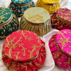 Op de afbeelding: Een verzameling kleurrijke, ronde stoffen pouches met ingewikkelde gouden borduursels. De pouches zijn verkrijgbaar in verschillende kleuren, waaronder rood, roze, groen en geel. Elke pouch heeft een bijpassende deksel en een ritssluiting. De tekst "THERAJASTHANIART" is zichtbaar.
