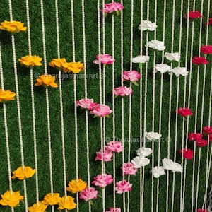 May include: A decorative backdrop with vertical strands of white beads against a green artificial grass background. The strands are decorated with yellow, pink, white, and red fabric flowers, creating a vibrant display.