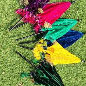 K&ouml;nnte beinhalten: F&uuml;nf bunte Samt-Schirme mit goldenen Blatt-Akzenten und Perlen-Quasten. Die Schirme sind f&auml;cherf&ouml;rmig auf einer gr&uuml;nen Grasfl&auml;che angeordnet.