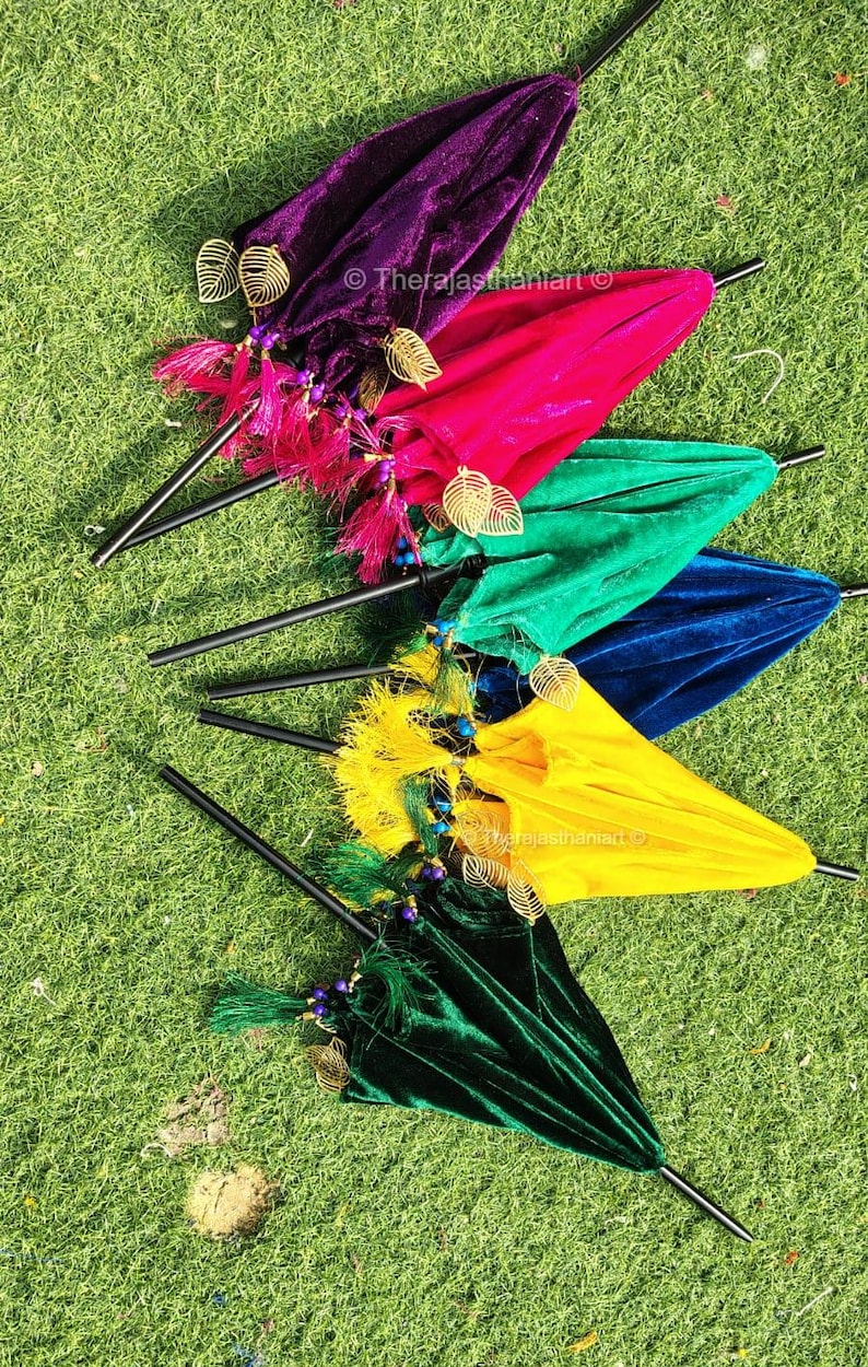 K&ouml;nnte beinhalten: F&uuml;nf bunte Samt-Schirme mit goldenen Blatt-Akzenten und Perlen-Quasten. Die Schirme sind f&auml;cherf&ouml;rmig auf einer gr&uuml;nen Grasfl&auml;che angeordnet.