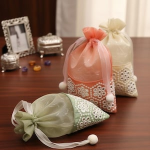 May include: Three decorative drawstring bags in the image. One is green, and two are peach and cream. The bags have white lace detailing and white pom-poms. The bags are on a dark wood surface.