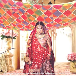 Puede incluir: Una novia con un lehenga rojo y dorado bordado se encuentra bajo un dosel colorido. El dosel está hecho de una tela estampada con un diseño geométrico. La novia lleva joyas tradicionales indias, incluyendo un aro de nariz, un collar y pulseras.