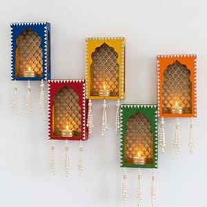 May include: Five rectangular wall-mounted candle holders in blue, yellow, orange, red, and green. Each features an arched cutout, a gold-toned lattice back panel, a small metallic tea light holder with a lit candle, and three hanging white bead tassels.