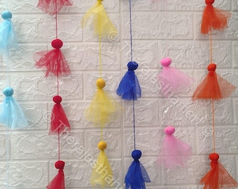 May include: A colourful garland of fabric tassels hanging from a white brick wall. The tassels are made of tulle and come in a variety of colours, including red, yellow, blue, pink, and light blue.