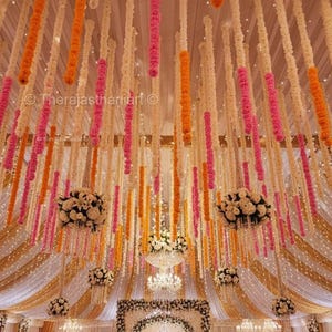 May include: A decorated event space with hanging floral garlands in shades of white, orange, and pink. The ceiling is adorned with cascading flower strands, creating a festive atmosphere. Chandeliers and draped fabric add to the elegant decor.