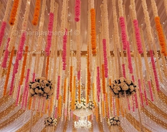 Könnte beinhalten: Veranstaltungsdekoration mit hängenden Blumengirlanden in Weiß, Orange und Pink. Die Decke ist mit kaskadenförmigen Blumensträngen geschmückt, die eine festliche Atmosphäre schaffen. Kronleuchter und drapierter Stoff tragen zur eleganten Einrichtung bei.
