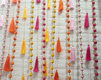 May include: A colourful garland of tassels and beads, featuring pink, orange, yellow, and gold accents. The garland is made of fabric and is strung together with gold thread.