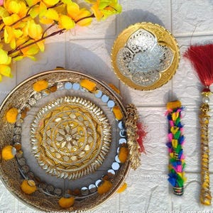 Può includere: Un piatto decorativo dorato e argentato con disegni intricati, un nappa rossa e un oggetto colorato con perline. Gli oggetti sono disposti su una superficie bianca con accenti floreali gialli e rosa.