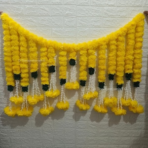 Puede incluir: Una guirnalda de flores de caléndula amarillas con detalles verdes y borlas blancas. La guirnalda está colgada en una pared de ladrillo blanco.