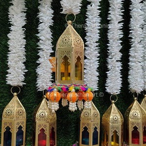 Può includere: Un set di sei lanterne in metallo dorato con pannelli in vetro colorati e disegni intricati. Ogni lanterna è adornata con un fiocco colorato e perline. Le lanterne sono appese a ghirlande di tessuto bianco.