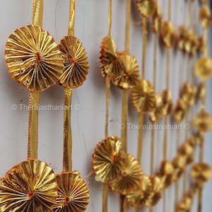 May include: Gold decorative garland featuring fan-shaped ornaments and vertical gold ribbons. The garland hangs against a white wall. The text "© Therajasthaniart" is visible.