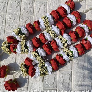 May include: Three floral garlands featuring alternating red rose and white flower clusters, interspersed with small yellow-green buds. Each garland is strung with a white beaded strand. The garlands are arranged on a textured white surface.