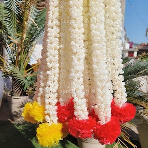 May include: A bunch of white flower garlands with red and yellow pom-poms at the bottom. The garlands are made of artificial flowers and are used for decoration.
