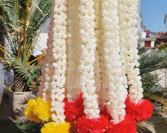 May include: A bunch of white flower garlands with red and yellow pom-poms at the bottom. The garlands are made of artificial flowers and are used for decoration.