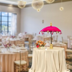 K&ouml;nnte beinhalten: Ein rosa Samt-Schirm mit goldenen Quasten und einem Holzgriff. Der Schirm steht auf einer wei&szlig;en Tischdecke auf einem Tisch in einem Raum mit wei&szlig;en W&auml;nden und einem gro&szlig;en Fenster.