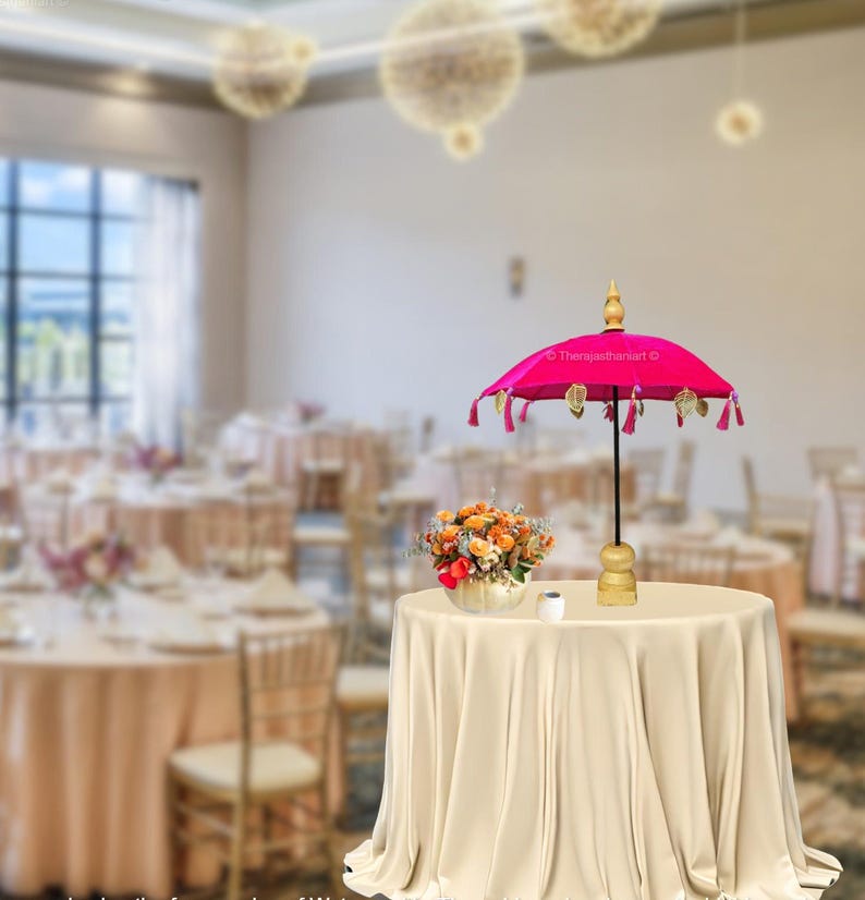 K&ouml;nnte beinhalten: Ein rosa Samt-Schirm mit goldenen Quasten und einem Holzgriff. Der Schirm steht auf einer wei&szlig;en Tischdecke auf einem Tisch in einem Raum mit wei&szlig;en W&auml;nden und einem gro&szlig;en Fenster.
