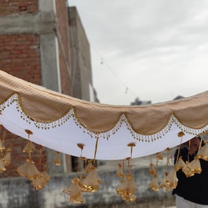 May include: A beige fabric canopy with a white lining and a beaded trim. The canopy is decorated with gold tassels hanging from the bottom edge.