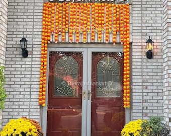 May include: A brick house with a red door decorated with yellow and orange marigold flower garlands. There are potted yellow mums on either side of the door.
