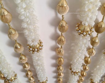 May include: Close-up of several strands of white floral garlands with gold and beige accents. Each strand features white flower clusters, gold-colored beads, and decorative gold and beige teardrop-shaped ornaments. The garlands are likely for festive or decorative purposes.