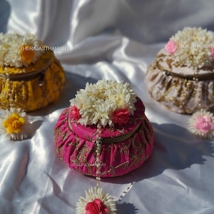 Puede incluir: Tres cajas redondas decorativas de tela en amarillo, rosa y rosa pálido, adornadas con bordados dorados y rematadas con arreglos florales blancos y rosas. Sobre un fondo de tela blanca.