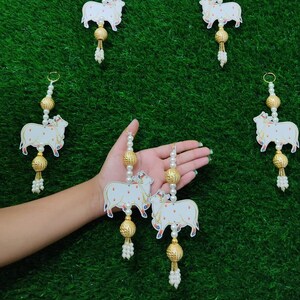 May include: Five white cow-shaped ornaments with gold accents and pearl beads hanging from a green background. The ornaments are strung with white beads and have a gold tassel at the bottom.