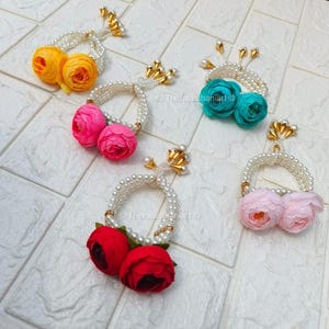 May include: Five floral garlands with pearl-like beads and gold accents. Each garland features two fabric flowers in various colours: yellow, pink, teal, red, and light pink. The garlands are arranged on a white tiled surface.