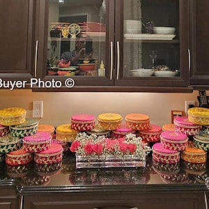 Op de afbeelding: Een stapel ronde, decoratieve dozen met gouden en roze versieringen. De dozen zijn in een piramidevorm gerangschikt en zijn versierd met ingewikkelde ontwerpen. De dozen worden waarschijnlijk gebruikt voor geschenken of cadeautjes.