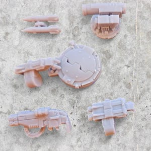 Può includere: Un set di cinque armi in miniatura stampate in 3D per un gioco da tavolo. Le armi sono in plastica grigia chiara e includono un lanciamissili, un cannone laser, una torretta, un lanciarazzi e un'arma piccola e rotonda.