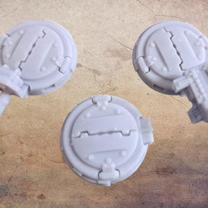 May include: Three light gray, circular miniature components with detailed mechanical designs. One has two cylindrical protrusions, another has a complex structure, and the third is a simple disc. These appear to be parts for a model or miniature.