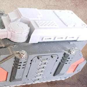 May include: A miniature armoured vehicle model featuring a dark grey chassis with orange accents on the side panels. The top section is a contrasting white, detailed with a circular hatch, a grid pattern, and various geometric moulded structures.