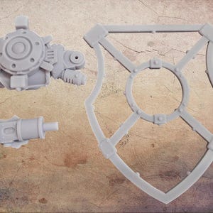 May include: Three unpainted, light gray plastic model parts. The parts include a mechanical arm, a cylindrical piece, and a shield-shaped frame with a central circle and cross-bracing. The pieces are likely for a miniature or model kit.