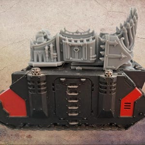 May include: A black and red miniature tank model featuring a side door with horizontal vents and two exhaust pipes. Mounted on top is a grey resin upgrade kit consisting of a central gate structure with a heart emblem and a spiked rear shield.