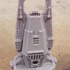 Puede incluir: Modelo en miniatura gris impreso en 3D de una estructura mecánica futurista. El modelo tiene un cuerpo central angular con paneles detallados, rejillas de ventilación y una serie de líneas verticales. Dos elementos rectangulares verticales se extienden desde la parte superior.