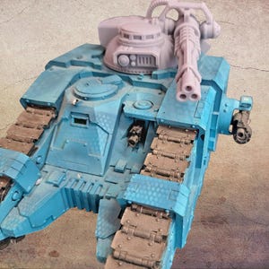 May include: A light blue model tank with a large gun turret and a multi-barreled gun. The tank has brown treads and various details, including hatches and a smaller gun on the side. The model is set against a textured background.