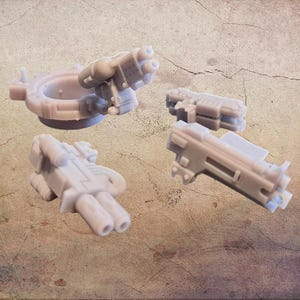 Peut inclure: Quatre modèles miniatures d'armes gris clair, dont une tourelle avec un canon à plusieurs canons et trois autres composants en forme de pistolet. Les objets sont disposés sur une surface brune texturée. Il s'agit de composants pour un modèle miniature.