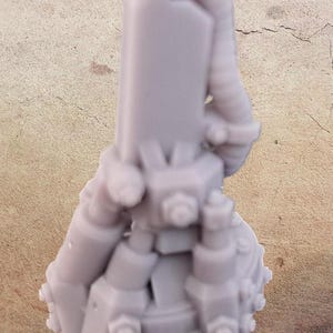 May include: A light gray, 3D-printed miniature, possibly for a tabletop game. The model features a cylindrical body with various attached mechanical components, including tubes and circular details. The base is round.