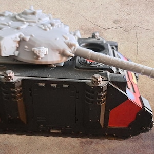 May include: A miniature tank model featuring a black and red chassis with metallic accents and a light gray, unpainted turret with a long barrel. The vehicle has a boxy, armored design with visible mechanical details and small skull-shaped ornaments.