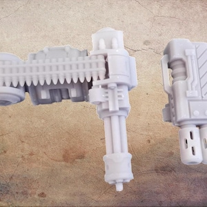 Puede incluir: Tres armas de juguete en miniatura de color gris claro se muestran sobre un fondo texturizado. El conjunto incluye una ametralladora Gatling, un arma de mano y un dispositivo rectangular. Estos modelos detallados son probablemente para aficionados o coleccionistas.